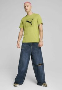 Camiseta verde de manga corta con un gráfico del logo negro de PUMA, combinada con unos vaqueros de mezclilla azul holgados, con desgastes y un corte relajado.
