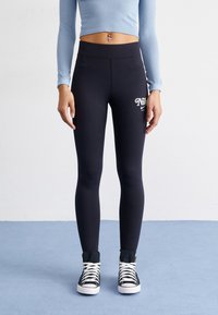 Leggings neri con dettagli del logo, vita alta, tessuto elasticizzato, abbinati a sneakers alte nere e un top cropped a maniche lunghe di colore azzurro chiaro.