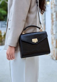 Schwarze Kunstleder-Handtasche im Krokodil-Design mit einer strukturierten Form, goldenen Beschlägen und einem Obergriff, getragen zu einem beigen Blazer mit Muster.