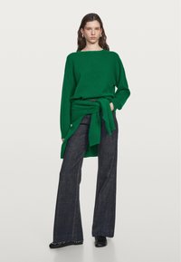 Grüner Strickpullover mit lockerer Passform, weitem Ärmeln und seitlicher Schleife, kombiniert mit hoch taillierten, dunkelblauen Jeans mit Schlag. Schwarze Schuhe.