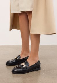 Kvinnliga ben i beige shorts och en tanfärgad kappa, kombinerat med svarta läderslippers med guldfäste på en betonggolv.