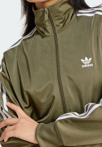 Olivově zelená bunda na zip s vysokým límcem, bílý tříproužkový design na rukávech a malým bílým logem na levé hrudi. Lesklá textura.