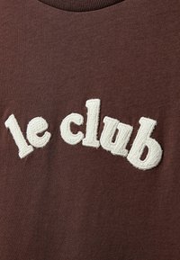Καφέ φούτερ με λευκή κεντημένη επιγραφή "le club." Το κείμενο έχει υφή. Στρογγυλή λαιμόκοψη με πλεκτό τελείωμα.