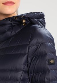 Marineblaue Steppjacke mit Kapuze, ausgestattet mit goldfarbenen Beschlägen und gestepptem Muster. Markenlabel auf dem Ärmel sichtbar. Glattes, glänzendes Material.