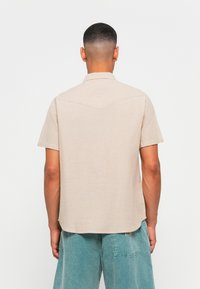 Short-sleeve, collared shirt in light beige fabric. Features a back yoke with a V-shaped seam. Worn with blue-green denim shorts.