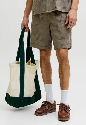 Mand iført teksturerede brune shorts og brune lædersko, der holder en stor cremefarvet og mørkegrøn lærredstaske i håndtagene.
