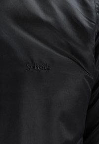 Gros plan sur un tissu noir avec un logo "Schott" délicatement brodé en fil noir assorti sur une surface textile lisse.