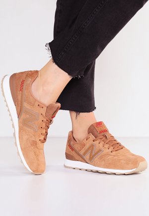 Personne portant des baskets New Balance en daim beige avec des surpiqûres rouges et des semelles blanches, associées à un jean noir raccourci effiloché.