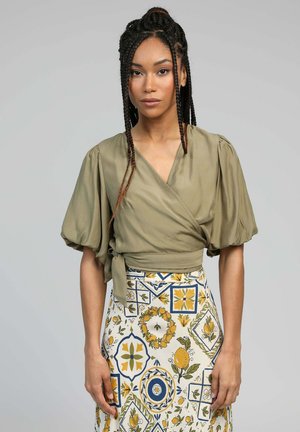Une femme avec des cheveux tressés porte un haut enroulé vert olive avec des manches bouffantes et une jupe à motifs présentant des designs floraux et fruitiers jaunes et bleus.
