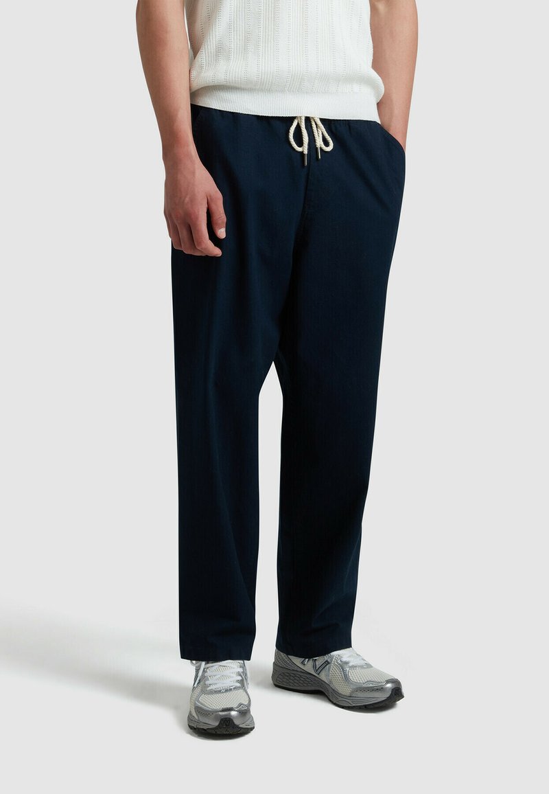 Marineblauwe, loszittende broek met een elastische tailleband en trekkoord. Gladde stoftextuur, zijzakken en een omgeslagen zoomdetail.