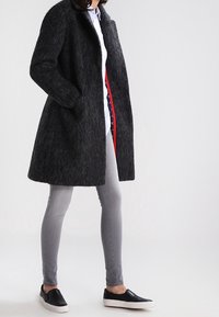 Abrigo de lana gris con corte recto, bolsillos laterales y forro rojo, combinado con pantalones vaqueros gris claro y zapatos sin cordones negros.