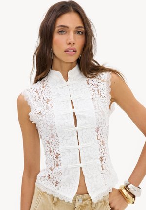 Kvinde iført en hvid blonde top uden ærmer med blomsterprint og traditionelle kinesiske knapper, kombineret med beige bukser og armbånd.