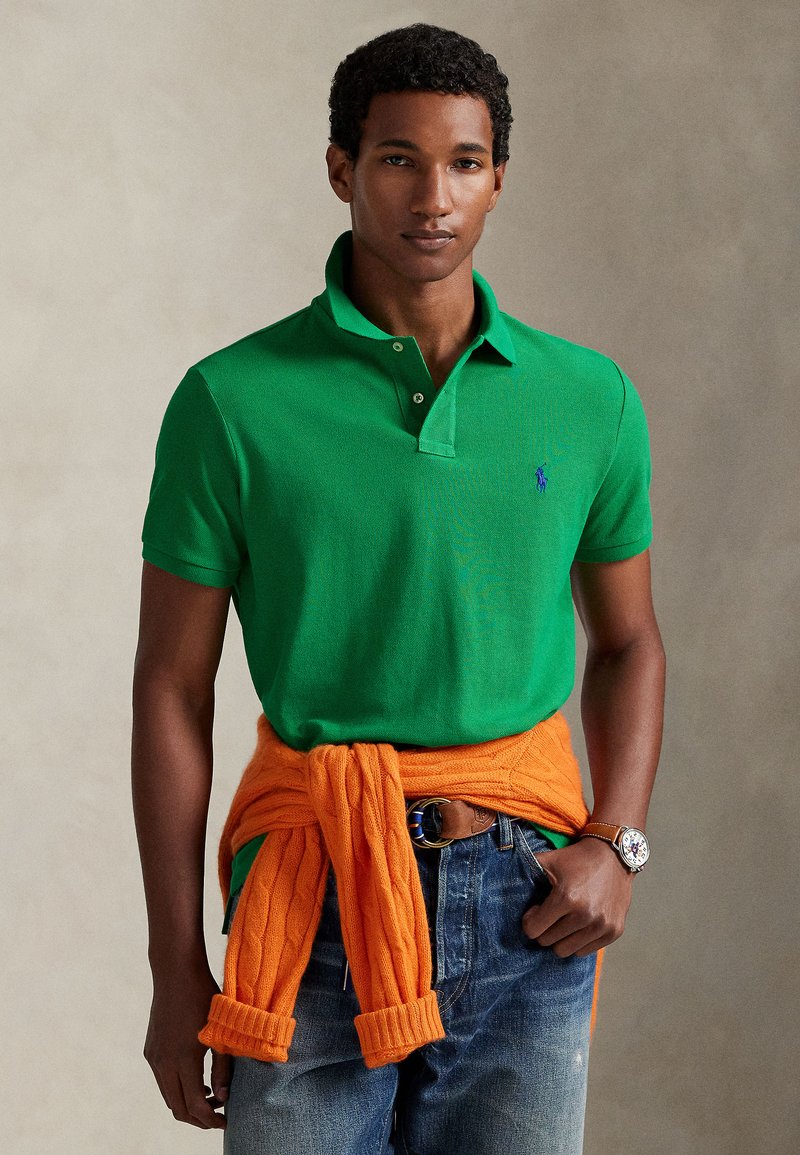 Πράσινο πόλο μπλουζάκι με κλασικό γιακά, σε συνδυασμό με πορτοκαλί πλεκτό πουλόβερ δεμένο στη μέση και μπλε τζιν. Κανονικό ρολόι στο χέρι.