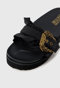 Sort slip-in sandal med volangkledd stropp og dekorativ spenne i gullfarge. Teksturert svart fotseng og såle.