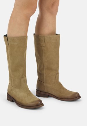 Gambe con stivali in camoscio color cuoio al ginocchio, con punte arrotondate e tacchi bassi impilati in legno su uno sfondo chiaro e uniforme.