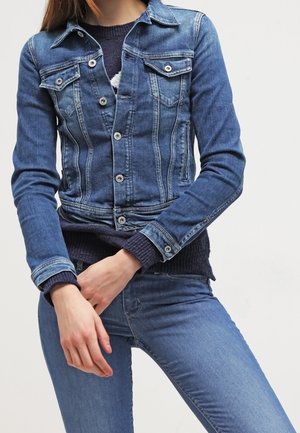 Personne portant une veste en jean bleue par-dessus un pull foncé et un jean bleu, debout les mains jointes devant.