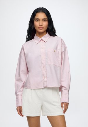 Un modèle aux longs cheveux foncés porte une chemise boutonnée rose pâle avec une poche poitrine et un short blanc, sur un fond clair et uni.