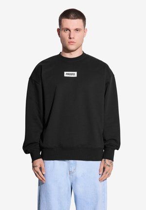 Czarny sweatshirt z bawełny, z okrągłym dekoltem, opadającymi ramionami, ściągaczami przy mankietach i białą prostokątną naszywką z logo na klatce piersiowej.