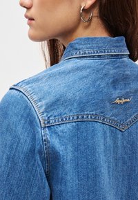 Μπουφάν τζιν σε ανοιχτό μπλε χρώμα με μυτερό γιακά, ορατές ραφές και μικρό λογότυπο στην πλάτη. Λεπτή υφή με κλασικό σχέδιο.