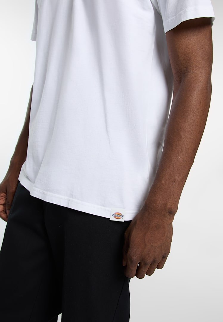 Uomo che indossa una t-shirt bianca semplice con una piccola etichetta con il logo Dickies vicino all'orlo, abbinata a pantaloni neri, su uno sfondo neutro.