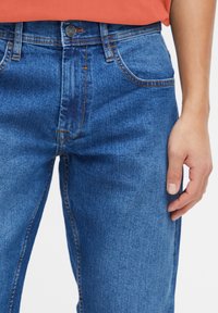 Jeans de denim azul con un corte recto, que cuentan con cinco bolsillos, costuras claras y un cierre de botón en la cintura. Mano reposando en el costado.