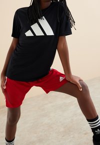 Camisa atlética de manga corta negra con gran logotipo blanco de Adidas, combinada con pantalones cortos rojos con rayas blancas y calcetines a rayas en blanco y negro.