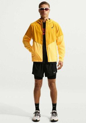 Żółta kurtka przeciwwiatrowa Nike z czarną dolną częścią, w zestawie z czarnymi szortami i białymi trampkami. Kurtka posiada kaptur i odblaskowe akcenty.