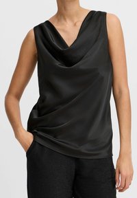 Top senza maniche in satin nero con scollo drappeggiato indossato da una persona con una mano nella tasca di pantaloni neri a trama.