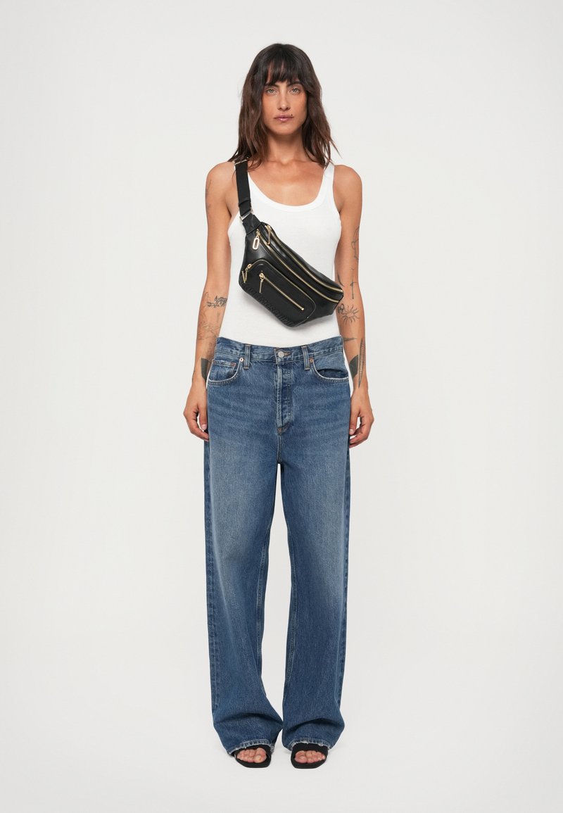 Schwarze Leder-Schultertasche mit mehreren Reißverschlussfächern, verziert mit goldfarbenen Beschlägen. Das Model trägt ein weißes Tanktop und lockere blaue Jeans.