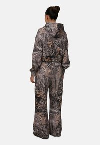Camo-patroon jumpsuit in gedempte bruin- en groentinten, met een capuchon, lange mouwen en ruime pijpen, gemaakt van zacht, duurzaam materiaal.