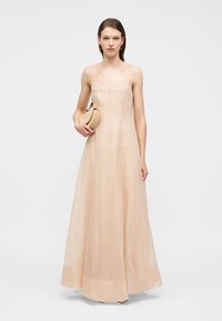 Γυναίκα που φοράει ένα μακρύ, αμάνικο, γυμνό φόρεμα, κρατώντας μια μικρή μπεζ τσάντα, στέκεται μπροστά σε απλό λευκό φόντο.