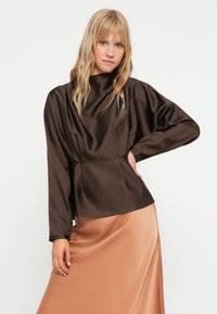 Bruine satijnen blouse met een hoge halslijn, lange mouwen en een aansluitende taille, gecombineerd met een vloeiende, lichtbruine satijnen rok. Gladde textuur.