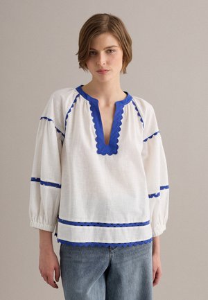Joven que lleva una blusa blanca con ribete azul con borde ondulado y mangas abullonadas hasta tres cuartos, combinada con unos vaqueros azules sobre un fondo liso.