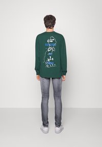 Camiseta verde de manga larga con un diseño de texto blanco y azul en la parte posterior, combinada con jeans gris claro y zapatillas blancas.