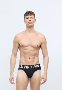 Crni pamučni bokserice s pojasom s logotipom "CALVIN KLEIN". Prikladan kroj, glatka tekstura, minimalno detaljiziranje. Model stoji na jednobojnoj pozadini.