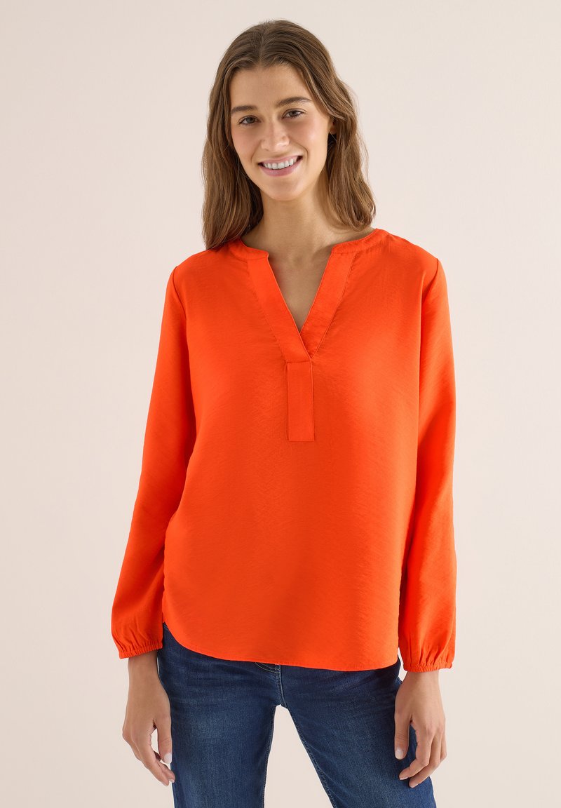 Jeune femme portant un chemisier orange vif à manches longues avec un col en V et un jean bleu, debout et souriante devant un fond uni.