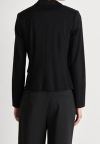 Zwarte blazer met lange mouwen, gestructureerde schoudernaad en een enkele achterventilatie. De stof lijkt soepel en vormgegeven voor een aansluitende silhouet.