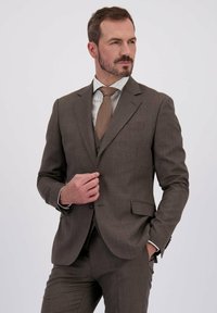 Man in een bruine pak met een gestreept overhemd en een bruine stropdas, die met één hand zijn jasje dichtknopt terwijl hij met de andere hand in zijn zak zit en naar links kijkt.