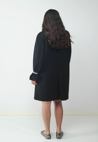 Schwarzes, übergroßes Kleid mit langen Ärmeln und einem Rückenschlitz, versehen mit silbernen Hardware-Details an den Manschetten. Glattes Material, knielanger Schnitt.