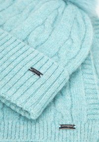 Jasnoniebieska dzianinowa czapka z wywiniętym ściągaczem, zdobiona wzorem warkocza. Metalowa tabliczka z nazwą marki zdobi czapkę. Miękka w dotyku tekstura i okrągły pompon na górze.