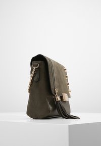 Handtasche aus olivgrünem Wildleder und Leder mit goldfarbenen Beschlägen, Quasten-Detail und dekorativen Nieten entlang der Klappe. Verfügt über einen Kettenriemen.