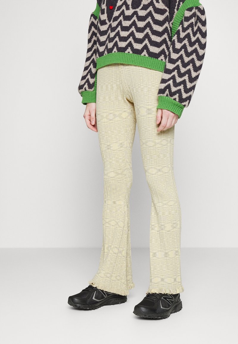 Pantalon évasé jaune clair avec un motif texturé et un ourlet à franges, associé à des chaussures de sport noires.