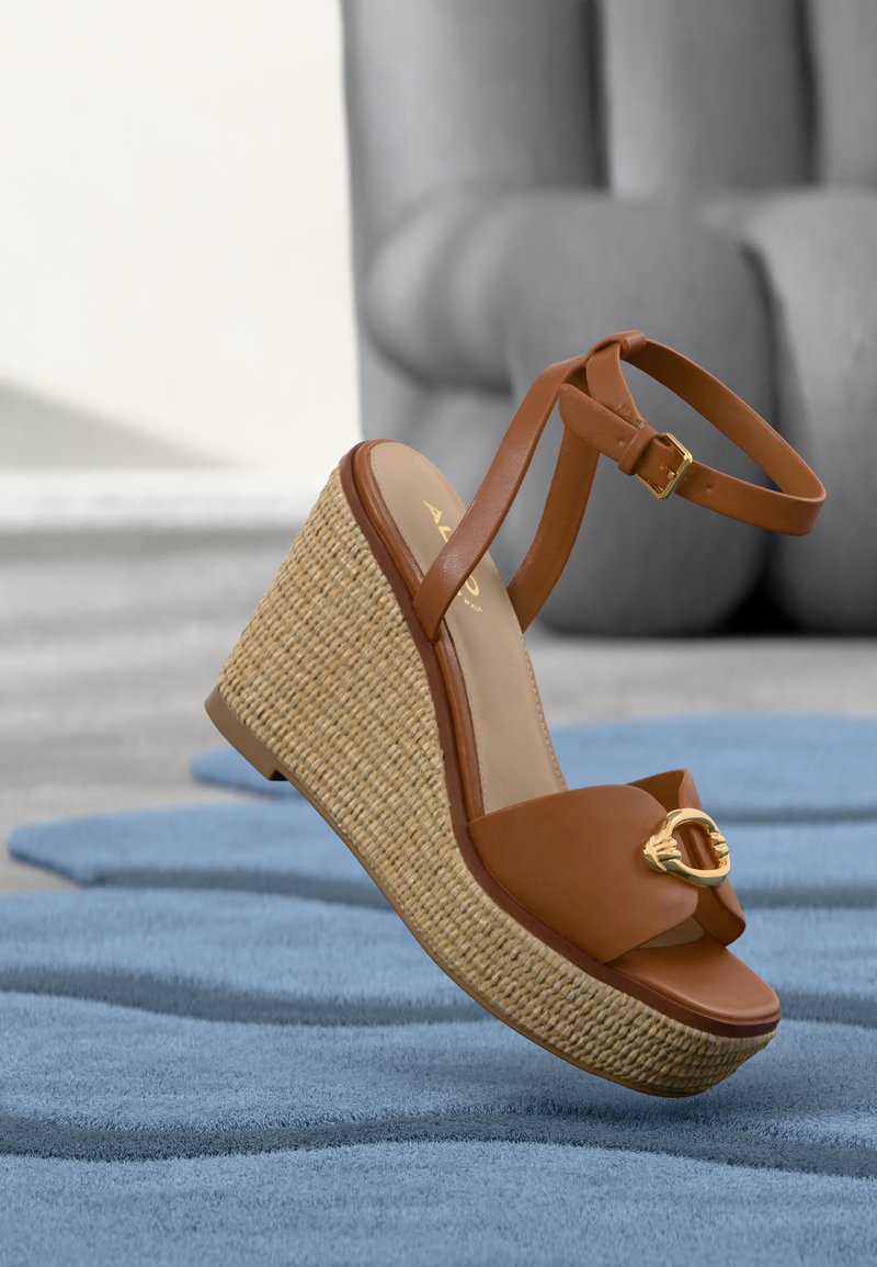 Bruine leren sleehak sandaal met gevlochten plateauzool en enkelriem, rustend op een blauw gestructureerd oppervlak binnenshuis.