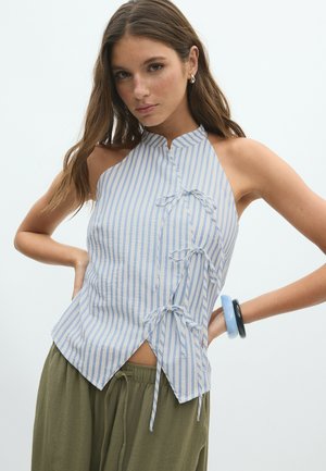 Jeune femme portant un haut dos nu rayé bleu avec des liens latéraux et un pantalon vert olive sur un fond blanc uni.