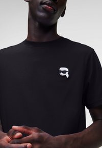 T-shirt nera in cotone con scollatura rotonda. Presenta una toppa di un personaggio dei cartoni animati bianco che indossa occhiali da sole sulla parte sinistra del petto.