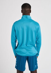 Camicia a maniche lunghe con colletto alto di un vivace ciano con una texture liscia. Abbinata a pantaloni corti blu scuro con strisce laterali e dettagli del logo.