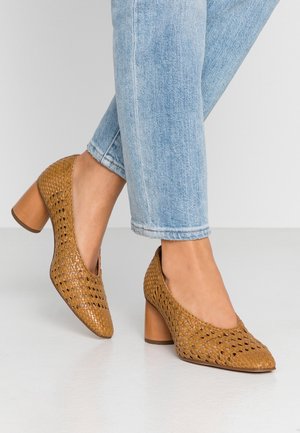 Hnědé pletené lodičky na vysokém podpatku s kulatou špičkou a výřezovým designem, spárované s světle modrými zkrácenými džíny na bílém povrchu.