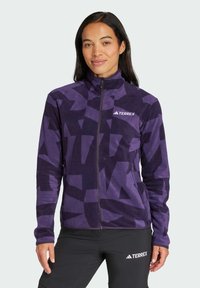 Chaqueta de forro polar morada con un patrón geométrico, cremallera completa, cuello alto y logo de Terrex en el pecho izquierdo. Textura suave, diseño ajustado.