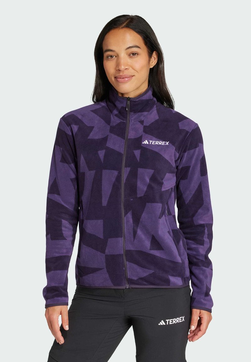 Chaqueta de forro polar morada con un patrón geométrico, cremallera completa, cuello alto y logo de Terrex en el pecho izquierdo. Textura suave, diseño ajustado.