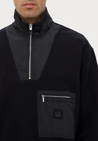 Pullover nero con colletto alto, dotato di zip frontale a metà e tasca frontale con zip e patch del marchio. Contrasto di tessuto texturizzato.